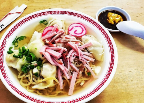 A bowl of noodles topped with ham, dumplings, green onions, and pink fish cake, served with a side of sauce.