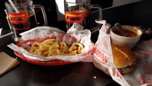 A table with curly fries, a sandwich, a bowl of chili, and two glasses of soda at a restaurant.