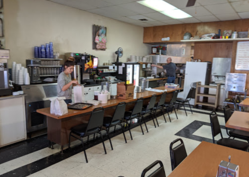 A diner interior with two staff members, a long table, chairs, and kitchen equipment in the background.