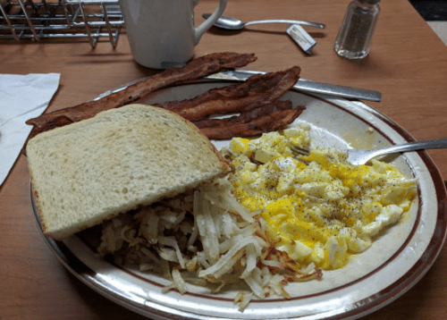 A plate of breakfast featuring scrambled eggs, crispy bacon, hash browns, and a slice of toast on a wooden table.