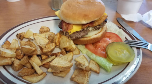A cheeseburger with bacon on a bun, served with lettuce, tomato, pickles, and a side of golden, crispy potato wedges.
