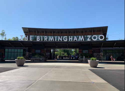 Entrance to the Birmingham Zoo, featuring a modern building and clear blue sky. Visitors are seen in the background.