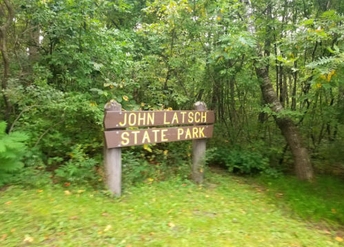 Wooden sign for John Latsch State Park surrounded by lush green trees and grass.