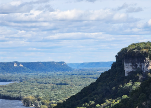 A scenic view of a river valley surrounded by lush greenery and cliffs under a partly cloudy sky.