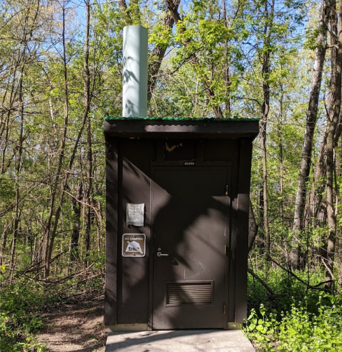 A small, dark brown building in a wooded area, featuring a door and a vent pipe on the roof.