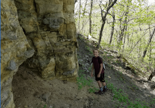A young boy stands near a rocky cliff in a wooded area, surrounded by greenery and trees.