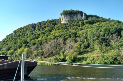 A scenic view of a lush green hillside with a rocky outcrop, seen from a river with a boat in the foreground.