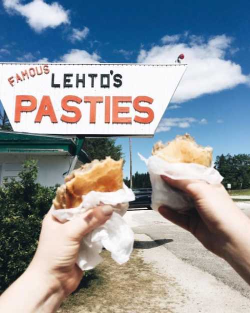 Two hands hold pasties in front of a sign that reads "Famous Lehto's Pasties" against a blue sky.