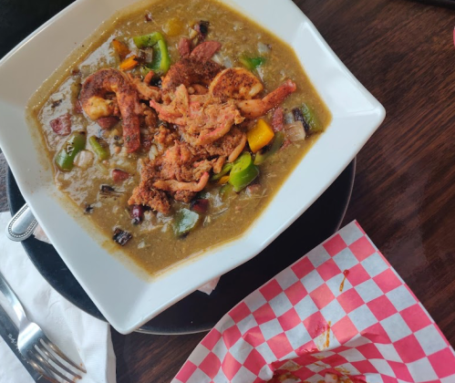 A bowl of shrimp gumbo with colorful bell peppers, served with a side of checkered paper for a takeout container.
