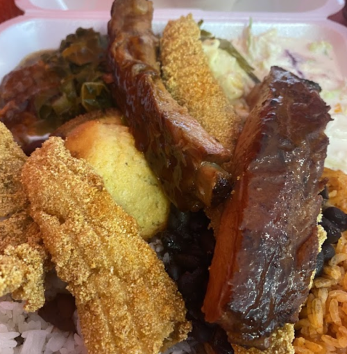 A close-up of a takeout container filled with rice, beans, fried fish, ribs, cornbread, and coleslaw.