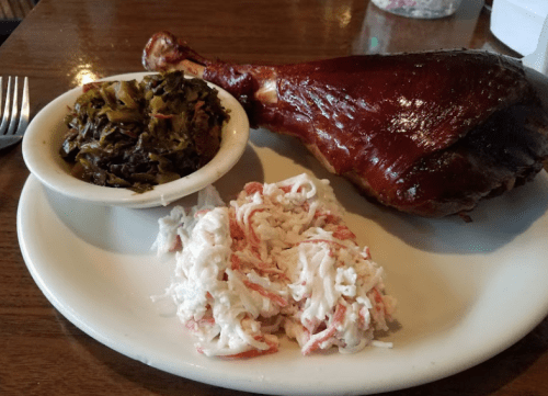 A plate with a smoked turkey leg, collard greens, and a serving of coleslaw on a wooden table.