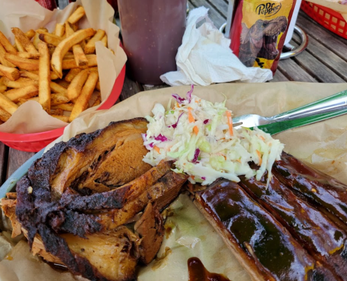A plate of barbecue ribs, coleslaw, and a side of fries, with a drink and napkin on a wooden table.