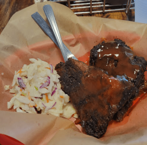 Two pieces of barbecued meat with sauce, served alongside a portion of coleslaw in a basket.