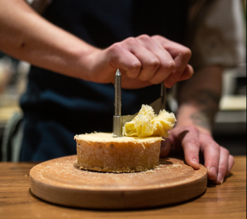 A chef uses a tool to scrape cheese from a round wheel onto a wooden board.