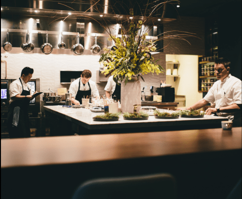 A modern kitchen with chefs preparing food, surrounded by pots and a large floral arrangement on the counter.