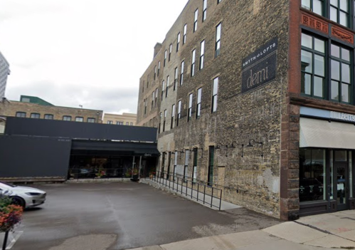 Exterior view of a brick building with a sign for "deli" and a parking area in front. Urban setting with cloudy sky.