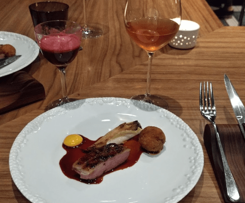 A gourmet plate featuring seared meat, sauce, a fried ball, and a yellow sauce, accompanied by two glasses of wine.