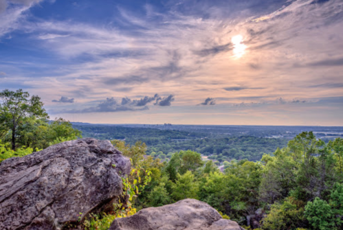 A scenic view from a rocky overlook, featuring a vibrant sky with clouds and a setting sun over a lush green landscape.