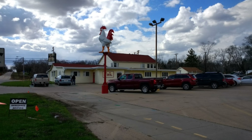 A roadside restaurant with a large rooster sign, surrounded by parked cars and a cloudy sky.