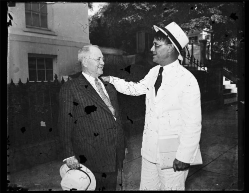 Two men in formal attire, one in a suit and the other in a white suit and hat, smiling and conversing outdoors.