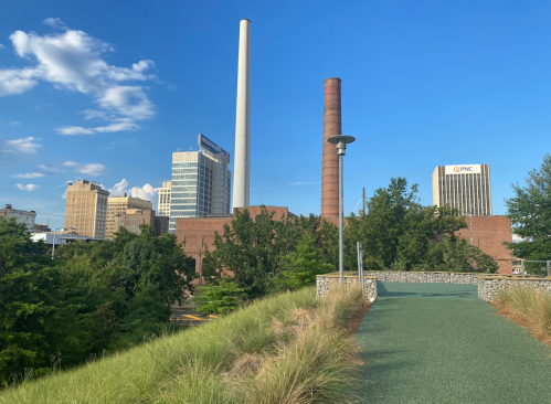 Cityscape featuring tall buildings, smokestacks, and greenery under a blue sky with scattered clouds.