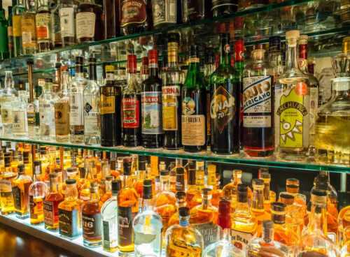 A well-stocked bar shelf displaying a variety of liquor bottles, including whiskey, gin, and liqueurs, illuminated warmly.