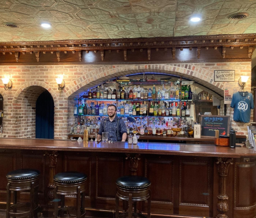 A bartender stands behind a wooden bar with a variety of bottles displayed on shelves in a cozy, warmly lit setting.