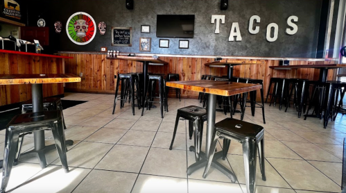 Interior of a taco restaurant featuring wooden tables, black chairs, and a "TACOS" sign on the wall.
