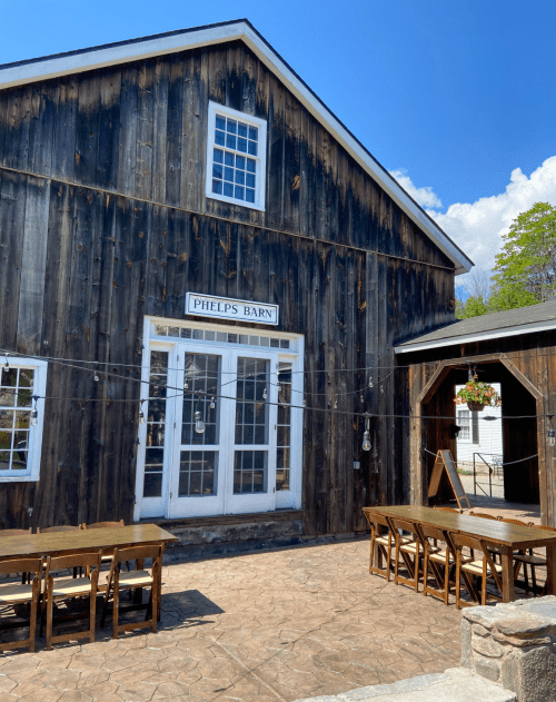 A rustic wooden barn with a sign reading "Phelps Barn," featuring outdoor seating and a clear blue sky.