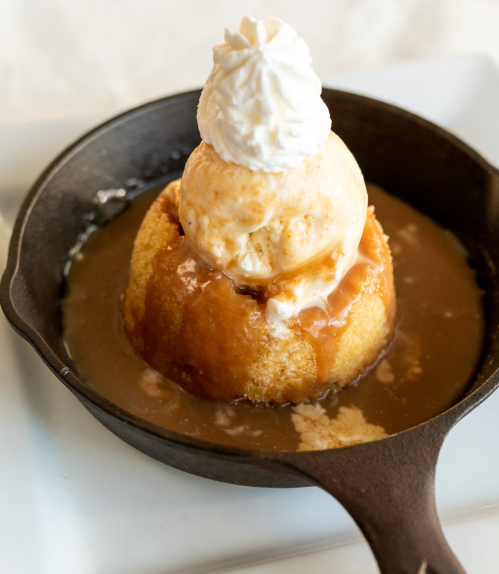 A warm dessert in a cast-iron skillet, featuring a cake topped with ice cream and drizzled with caramel sauce.