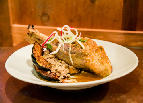 A plated dish featuring a roasted meat leg, garnished with green onions, served with grains and roasted vegetables.
