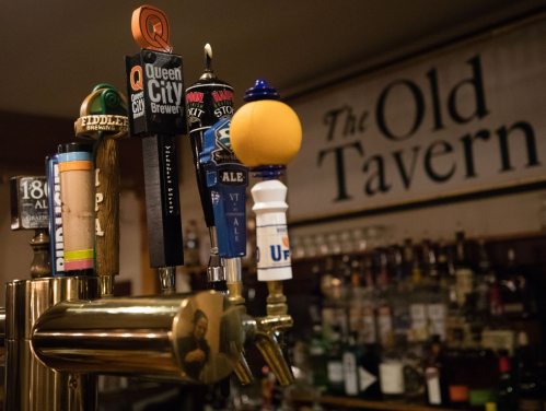 A close-up of beer taps at a bar, with various colorful tap handles and a sign reading "The Old Tavern" in the background.