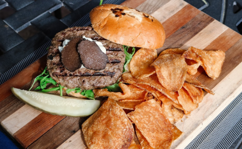 A grilled burger with lettuce, two slices of cheese, served with potato chips and a pickle on a wooden board.