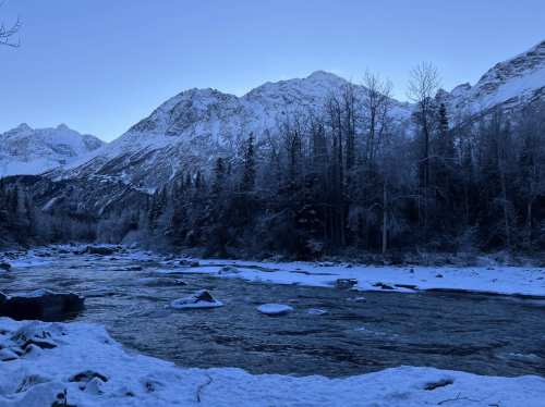 A serene winter landscape featuring a river, snow-covered ground, and mountains under a clear blue sky.