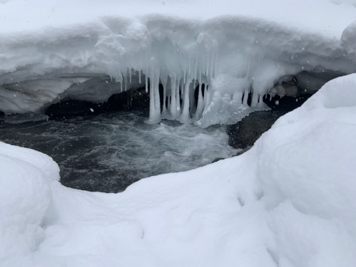 A snowy landscape with icicles hanging over a flowing stream, surrounded by white snow.