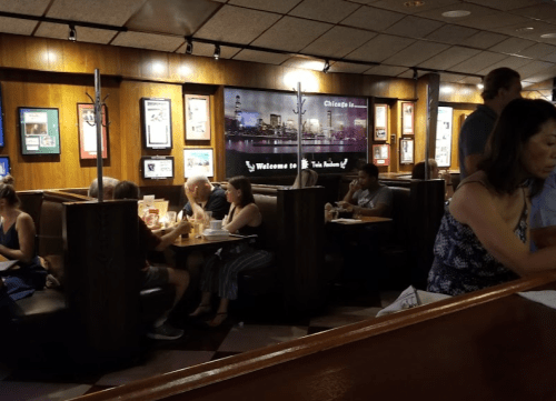 A busy restaurant interior with diners in booths and a woman serving at the counter. Warm lighting and decor visible.