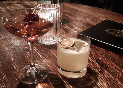 A cocktail glass with a cherry and a short glass with a light drink, both on a wooden table.