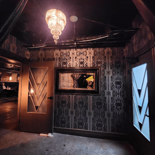 A stylish interior with patterned walls, a chandelier, and two doors, one illuminated, reflecting a vintage aesthetic.