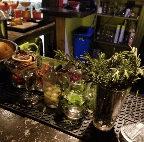 A bar counter with various glasses filled with herbs, fruits, and garnishes, alongside cocktail ingredients in the background.