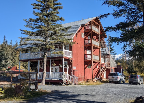 A red multi-story building surrounded by trees, with balconies and a gravel driveway under a clear blue sky.