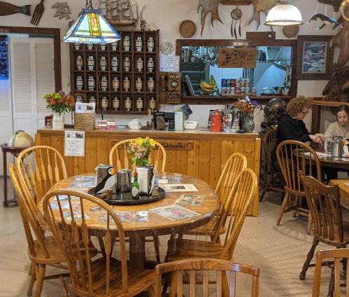 Cozy café interior with wooden tables, chairs, and a counter displaying jars and snacks, featuring two people at a table.