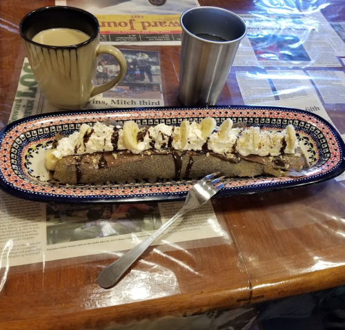 A plate with a chocolate-drizzled banana crepe, topped with whipped cream, alongside two cups of coffee.