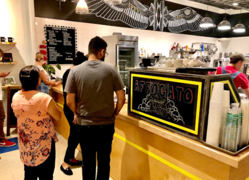 A busy café scene with customers waiting in line at the counter, featuring a chalkboard menu and coffee equipment.
