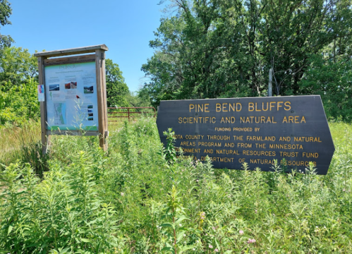 Sign for Pine Bend Bluffs Scientific and Natural Area, surrounded by greenery and trees under a clear blue sky.