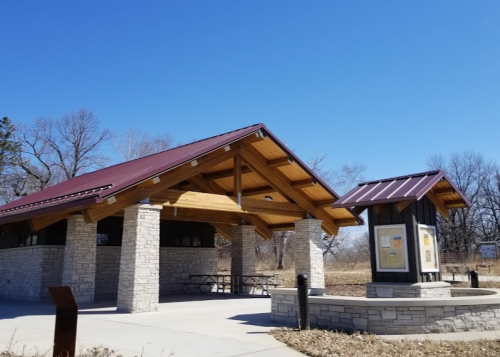 A pavilion with a wooden roof and stone columns, set against a clear blue sky, with picnic tables underneath.