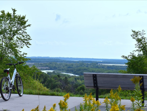 A bike rests beside a bench overlooking a lush green landscape and winding river under a cloudy sky.