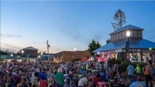 A large crowd gathers at an outdoor event near a pavilion during sunset, with colorful lights and festive atmosphere.