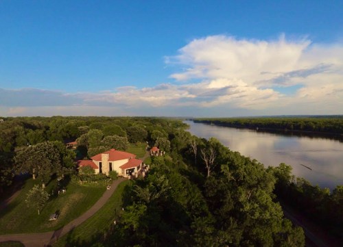 Aerial view of a river bordered by lush greenery and a house with a red roof under a blue sky with clouds.