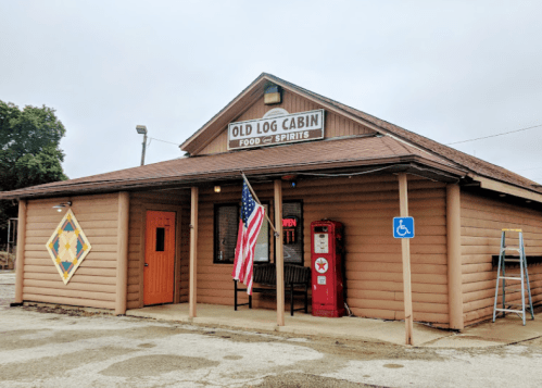 A rustic building with a sign reading "Old Log Cabin Food & Spirits," featuring an American flag and a handicapped-accessible entrance.