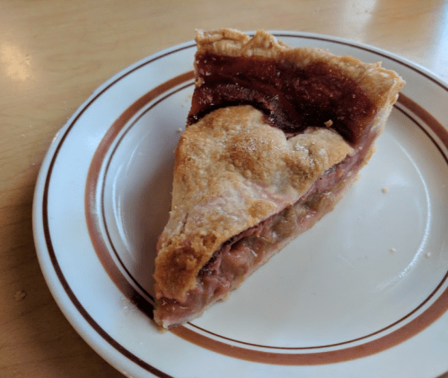 A slice of pie with a flaky crust and a rich, red filling on a white plate with a brown rim.